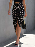 A woman wearing a high waist, midi length, polka dot skirt with a drawstring waist and a slit in the front. She is also carrying a black handbag and wearing black strappy sandals.
