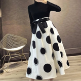 Person wearing a black turtleneck and white skirt with black polka dots in an indoor setting.